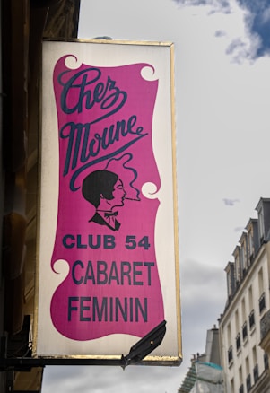 A vertical sign displays the name 'Chez Moune' with an elegant script font against a pink background. Below, the text 'Club 54 Cabaret Feminin' is written in bold, black letters. An illustration of a woman's profile, styled in a vintage silhouette, is depicted as well. The sign is mounted on the exterior of a building against a backdrop of clouds and neighboring structures.