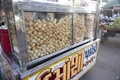 A street food cart displaying a large quantity of crispy, round snacks inside a glass case. The cart is adorned with colorful signage in a local script, and there are bags of snacks stored beside the main display. In the background, a marketplace is visible with various fruits and people.