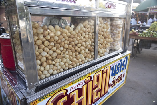 A street food cart displaying a large quantity of crispy, round snacks inside a glass case. The cart is adorned with colorful signage in a local script, and there are bags of snacks stored beside the main display. In the background, a marketplace is visible with various fruits and people.
