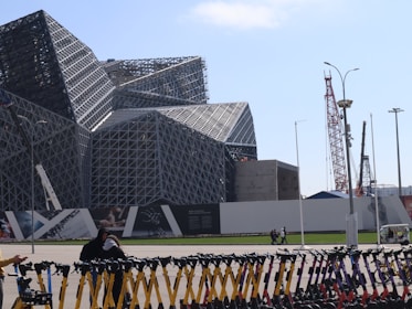 A large, modern building with a complex geometric pattern and metallic framework dominates the scene. In the foreground, multiple rows of neatly arranged electric scooters are visible. Several cranes are positioned in the background, indicating nearby construction activities. A few people are walking or standing around on the pavement.