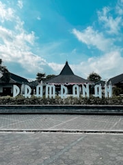 A building with a triangular roof surrounded by lush greenery and a clear blue sky. The word 'PRAMBANAN' is prominently displayed in large white letters in front of the structure, likely indicating a location or landmark. The scene is well-lit with natural daylight, enhancing the vivid colors of the sky and foliage.