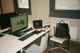 A modern workspace with two computer monitors, a laptop, and various personal items on a white desk. The main monitor displays a green map, while the laptop screen is partially visible. Accessories such as glasses, a smartphone, a pair of headphones, and a black leather backpack placed on a chair are also present. The setting includes two large windows and minimalist furniture.