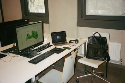 A modern workspace with two computer monitors, a laptop, and various personal items on a white desk. The main monitor displays a green map, while the laptop screen is partially visible. Accessories such as glasses, a smartphone, a pair of headphones, and a black leather backpack placed on a chair are also present. The setting includes two large windows and minimalist furniture.