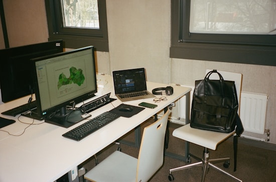 A modern workspace with two computer monitors, a laptop, and various personal items on a white desk. The main monitor displays a green map, while the laptop screen is partially visible. Accessories such as glasses, a smartphone, a pair of headphones, and a black leather backpack placed on a chair are also present. The setting includes two large windows and minimalist furniture.
