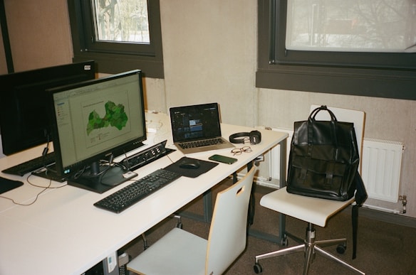 A modern workspace with two computer monitors, a laptop, and various personal items on a white desk. The main monitor displays a green map, while the laptop screen is partially visible. Accessories such as glasses, a smartphone, a pair of headphones, and a black leather backpack placed on a chair are also present. The setting includes two large windows and minimalist furniture.