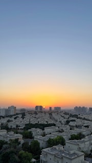 A cityscape at sunset featuring clusters of white residential buildings surrounded by greenery. Tall modern buildings are in the background, with the sun setting near the horizon, casting a warm orange glow in the sky that fades into a clear blue above.