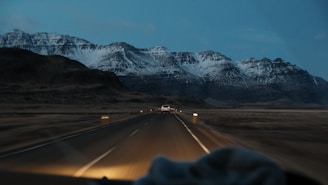 A winding road leads through a vast, barren landscape with towering snow-capped mountains in the background under a dim, overcast sky. A vehicle is visible on the road, heading towards the mountains, creating a sense of journey and adventure.
