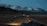 A winding road leads through a vast, barren landscape with towering snow-capped mountains in the background under a dim, overcast sky. A vehicle is visible on the road, heading towards the mountains, creating a sense of journey and adventure.
