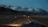 A winding road leads through a vast, barren landscape with towering snow-capped mountains in the background under a dim, overcast sky. A vehicle is visible on the road, heading towards the mountains, creating a sense of journey and adventure.