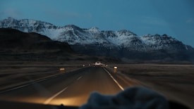 A winding road leads through a vast, barren landscape with towering snow-capped mountains in the background under a dim, overcast sky. A vehicle is visible on the road, heading towards the mountains, creating a sense of journey and adventure.
