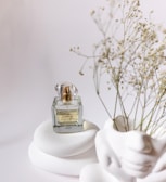 A clear glass perfume bottle with gold accents sits atop smooth, white, rounded stones. Delicate dried flowers are positioned to the right, adding a natural element against the minimalist white background. The bottle is labeled with elegant script.