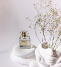 A clear glass perfume bottle with gold accents sits atop smooth, white, rounded stones. Delicate dried flowers are positioned to the right, adding a natural element against the minimalist white background. The bottle is labeled with elegant script.