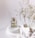 A clear glass perfume bottle with gold accents sits atop smooth, white, rounded stones. Delicate dried flowers are positioned to the right, adding a natural element against the minimalist white background. The bottle is labeled with elegant script.