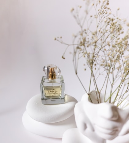 A clear glass perfume bottle with gold accents sits atop smooth, white, rounded stones. Delicate dried flowers are positioned to the right, adding a natural element against the minimalist white background. The bottle is labeled with elegant script.