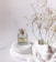 A clear glass perfume bottle with gold accents sits atop smooth, white, rounded stones. Delicate dried flowers are positioned to the right, adding a natural element against the minimalist white background. The bottle is labeled with elegant script.