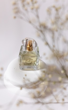 A glass perfume bottle with a gold atomizer is placed on a white stone surface. In the background, there are soft-focus branches with small white flowers, giving a delicate and elegant atmosphere.