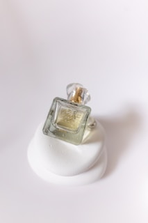 A small, square glass perfume bottle with a transparent cap, partially filled with a light yellow liquid, resting on a white, abstract, layered surface.