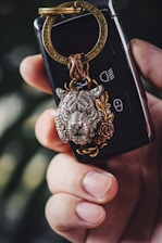 A close-up of a hand holding a set of car keys with a decorative keychain featuring an intricately detailed lion's head. The lion's head appears to be made of metal with a bronze and silver color finish, and is adorned with a rose design. The key fob is black with visible buttons and is secured by a gold-colored keyring.