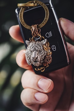 A close-up of a hand holding a set of car keys with a decorative keychain featuring an intricately detailed lion's head. The lion's head appears to be made of metal with a bronze and silver color finish, and is adorned with a rose design. The key fob is black with visible buttons and is secured by a gold-colored keyring.