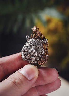 A hand holding a small, intricately crafted pendant featuring a detailed wolf's head surrounded by ornate floral designs. The pendant appears metallic with a mix of silver and gold tones, highlighting its craftsmanship.