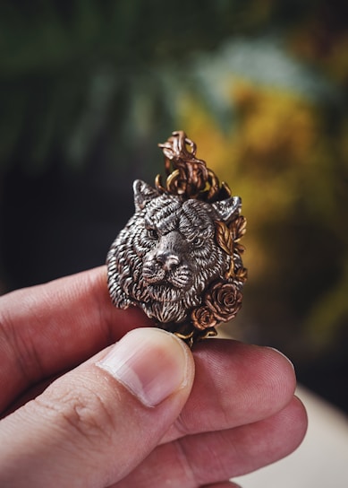 A hand holding a small, intricately crafted pendant featuring a detailed wolf's head surrounded by ornate floral designs. The pendant appears metallic with a mix of silver and gold tones, highlighting its craftsmanship.