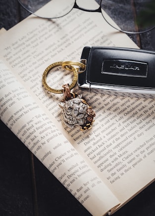An open book with a key fob resting on it. The key fob has a detailed lion head keychain attached. Above the book, a pair of eyeglasses is visible, partially in frame, suggesting a sense of reading or study.