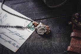 A rustic wooden table surface holds a vintage postcard with handwritten script. Nearby, a detailed necklace with a lion's head pendant and a pocket watch with a leather strap add to the antique aesthetic.