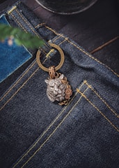 A close-up view of a pair of dark blue denim jeans featuring prominent yellow stitching. Attached to the jeans is a decorative metal keychain with a detailed lion head design, accentuated by floral motifs. The background includes a blurred element that resembles a plant or tree branch.