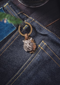 A close-up view of a pair of dark blue denim jeans featuring prominent yellow stitching. Attached to the jeans is a decorative metal keychain with a detailed lion head design, accentuated by floral motifs. The background includes a blurred element that resembles a plant or tree branch.