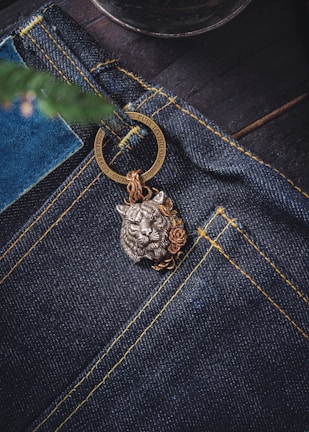 A close-up view of a pair of dark blue denim jeans featuring prominent yellow stitching. Attached to the jeans is a decorative metal keychain with a detailed lion head design, accentuated by floral motifs. The background includes a blurred element that resembles a plant or tree branch.