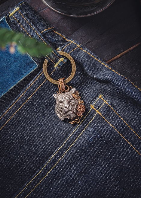 A close-up view of a pair of dark blue denim jeans featuring prominent yellow stitching. Attached to the jeans is a decorative metal keychain with a detailed lion head design, accentuated by floral motifs. The background includes a blurred element that resembles a plant or tree branch.