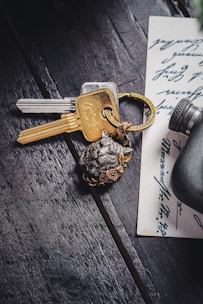 A set of keys with a metallic keychain featuring an animal face and floral design lies on a dark wooden surface. Beside the keys is a handwritten note with cursive writing, partially visible. A small cylindrical object is near the edge of the note.