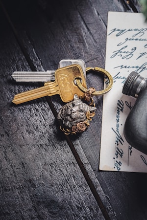 A set of keys with a metallic keychain featuring an animal face and floral design lies on a dark wooden surface. Beside the keys is a handwritten note with cursive writing, partially visible. A small cylindrical object is near the edge of the note.