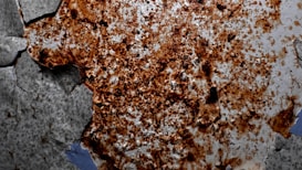 A large area of rust covers a surface, with the corrosion creating irregular patterns of orange, brown, and black discoloration. The surrounding area is a lighter gray, showing signs of old paint or a weathered material with cracks and peeling sections. The texture appears rough and uneven, indicating significant wear and deterioration.