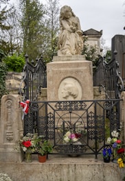 A grave is adorned with a statue of a seated woman holding a lyre. The gravestone includes an engraved portrait and is surrounded by intricate ironwork fencing. Flowers and small potted plants are placed at the base, adding a touch of color.