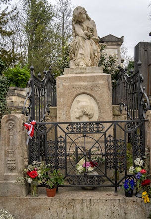 A grave is adorned with a statue of a seated woman holding a lyre. The gravestone includes an engraved portrait and is surrounded by intricate ironwork fencing. Flowers and small potted plants are placed at the base, adding a touch of color.