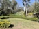 A landscaped garden area with well-maintained lawns and various types of green bushes and plants. The centerpiece is a large floral clock surrounded by a small fence. Tall trees and dense foliage provide a lush backdrop.