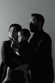 A family of three is captured in a side profile shot. The adults are looking in the same direction, with a child in the arms of one of the adults. They appear to be indoors, and the lighting creates a dramatic, shadowed effect on their faces.