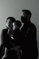 A family of three is captured in a side profile shot. The adults are looking in the same direction, with a child in the arms of one of the adults. They appear to be indoors, and the lighting creates a dramatic, shadowed effect on their faces.