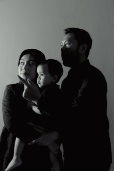 A family of three is captured in a side profile shot. The adults are looking in the same direction, with a child in the arms of one of the adults. They appear to be indoors, and the lighting creates a dramatic, shadowed effect on their faces.