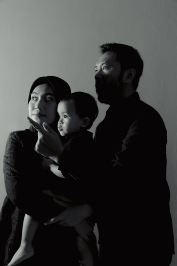 A family of three is captured in a side profile shot. The adults are looking in the same direction, with a child in the arms of one of the adults. They appear to be indoors, and the lighting creates a dramatic, shadowed effect on their faces.