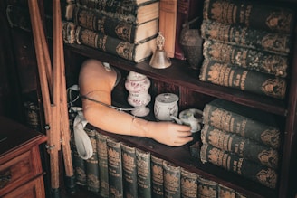 A wooden bookshelf filled with old, worn encyclopedias and other antique books. A small porcelain teacup set is placed on one of the shelves next to a vintage prosthetic arm. A small metal bell is also present, along with some walking canes leaning on the shelf.