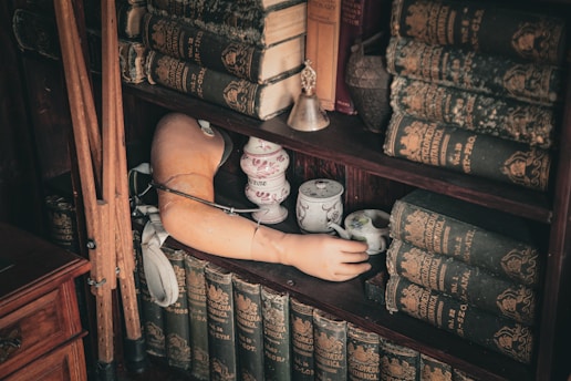 A wooden bookshelf filled with old, worn encyclopedias and other antique books. A small porcelain teacup set is placed on one of the shelves next to a vintage prosthetic arm. A small metal bell is also present, along with some walking canes leaning on the shelf.