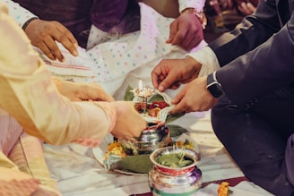 A group of people participate in a traditional ceremony, placing offerings into a metal pot surrounded by leaves and flowers. The participants are wearing formal attire, and one person is holding a spoon, possibly performing a ritual. The setting has a festive ambiance with colorful elements.