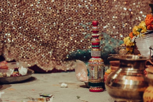 A richly decorated ceremonial setup with a bejeweled, ornate bottle standing upright surrounded by traditional brass utensils, vibrant flowers, and other ceremonial items. The background is a shimmering, intricately patterned fabric, adding a festive and cultural atmosphere to the scene.