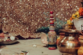 A richly decorated ceremonial setup with a bejeweled, ornate bottle standing upright surrounded by traditional brass utensils, vibrant flowers, and other ceremonial items. The background is a shimmering, intricately patterned fabric, adding a festive and cultural atmosphere to the scene.