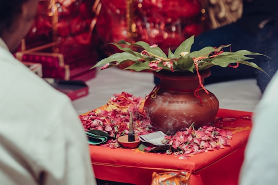 A traditional setup featuring a clay pot adorned with green leaves, placed on a red cloth. Surrounding the pot are scattered flower petals and a lit incense stick. The background includes vibrant red and gold decorations, suggesting a ceremonial or festive context.