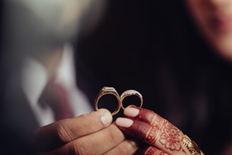 Hands with intricate mehndi designs hold two gold rings, one with diamond embellishments. The soft focus creates an intimate atmosphere.