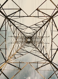 A metal transmission tower viewed from directly underneath, emphasizing its intricate network of beams and wires. The structure forms geometric patterns against a cloudy sky, creating a sense of depth and vertical perspective.