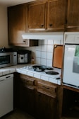 A cozy kitchen corner with wooden cabinets, a small stove with two black electric burners, and a black frying pan on one burner. A microwave and coffee maker are placed on the counter, and there are white tiled countertops with a backsplash. A cutting board is propped against the wall beside the stove.